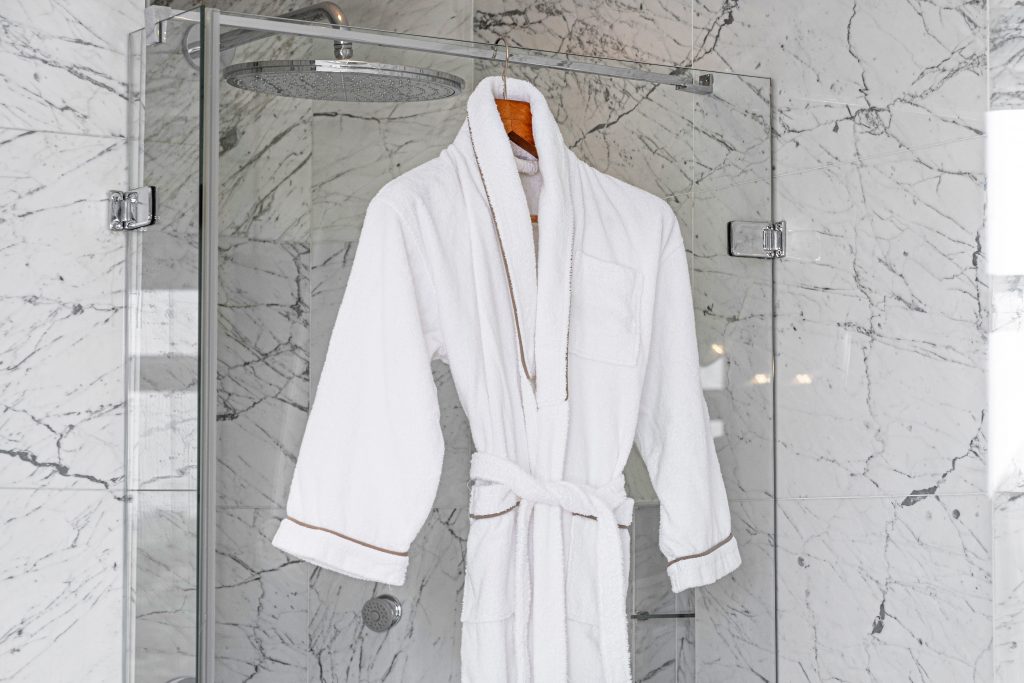 White bathrobe hanging on door of a modern white marble bathroom.